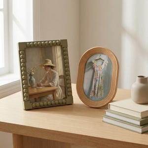 May include: A rectangular sage green picture frame with a beaded border containing a painting of a person in a hat at a table, and an oval light wood frame with a ribbed texture containing a painting of a floral dress on a hanger.