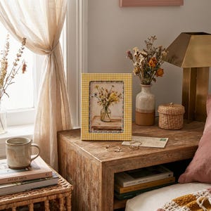 May include: A yellow gingham-patterned picture frame with a floral print. The frame sits on a wooden nightstand, next to a vase of dried flowers and a small woven basket. A mug and books are also visible.