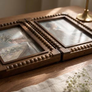 May include: A hinged double picture frame made of dark brown wood with a decorative beaded border. The frame holds two separate prints, one depicting hands holding a bouquet of flowers and the other showing a person in a light-colored garment.
