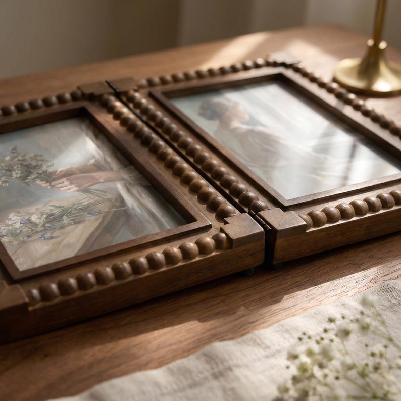 May include: A hinged double picture frame made of dark brown wood with a decorative beaded border. The frame holds two separate prints, one depicting hands holding a bouquet of flowers and the other showing a person in a light-colored garment.