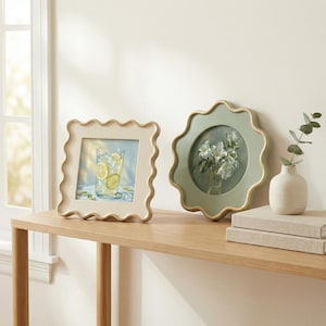 May include: A square cream-colored wavy frame containing a print of a lemon drink and a round sage green wavy frame containing a floral print, positioned next to a small white ceramic vase with a green sprig resting on two stacked books.