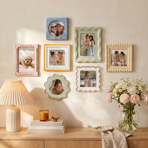 May include: A collection of seven wall-mounted picture frames in various shapes and colors, including square, rectangular, and scalloped designs in blue, pink, orange, green, and cream, surrounding a table with a lamp and a vase of flowers.