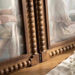 May include: A hinged double picture frame constructed from dark brown wood, featuring a decorative beaded trim along the edges. The frame holds two separate paintings depicting figures in white clothing, connected by a small metal hinge.