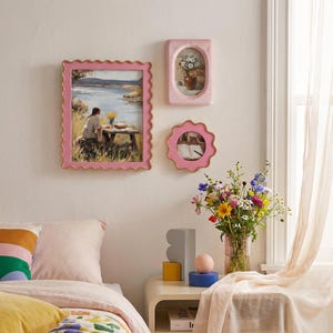 May include: A collection of pink picture frames with gold trim, each holding different artwork. One frame has a scalloped edge and displays a landscape painting. Another is oval-shaped, and the third is a scalloped circle.