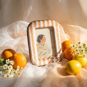 May include: A rectangular picture frame with a white and tan vertical striped pattern. The frame holds a portrait of a person wrapped in a light-colored shawl. The frame is surrounded by fresh oranges, lemons, and small white daisy-like flowers.