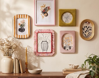 Peut inclure: Un ensemble de six cadres photo variés aux motifs roses, jaunes, dorés et rayés, présentant des illustrations botaniques et des portraits. En dessous, une console en bois supporte un vase en céramique avec des fleurs séchées, des livres, une bougie et un bol.