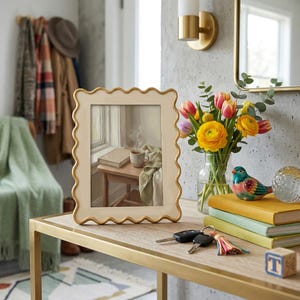 May include: A decorative table with a gold frame and a light wood top. A framed picture with a scalloped edge sits on the table. A vase of flowers, books, keys, and a small bird figurine are also on the table.