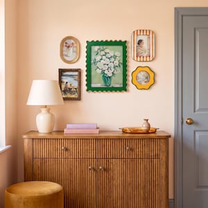 May include: A wooden sideboard with a fluted front, a cream-colored table lamp, and a stack of two books. Above, a gallery wall features a large green scalloped frame with a floral print, two rectangular frames, and a yellow flower-shaped frame.