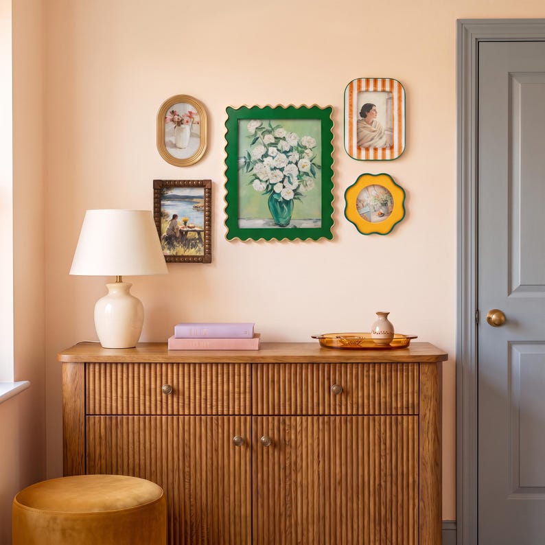 May include: A wooden sideboard with a fluted front, a cream-colored table lamp, and a stack of two books. Above, a gallery wall features a large green scalloped frame with a floral print, two rectangular frames, and a yellow flower-shaped frame.