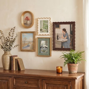 May include: A gallery wall arrangement featuring five framed art prints in various shapes and sizes, including an oval gold frame, a scalloped white frame, two rectangular gold beaded frames, and a large dark brown scalloped frame.