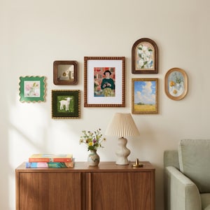 May include: A gallery wall arrangement featuring six framed artworks of varying shapes and sizes, including rectangular, square, and arched frames. Below, a wooden sideboard holds two stacked books, a small ceramic vase with flowers, and a lamp.