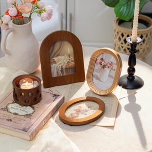 May include: A collection of decorative items on a table, including wooden picture frames, a candle holder with a lit candle, a vase with flowers, and a book. The frames display images of flowers and a candlelit scene.