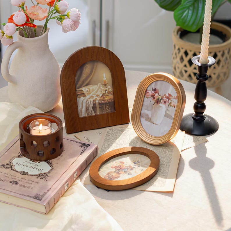 May include: A collection of decorative items on a table, including wooden picture frames, a candle holder with a lit candle, a vase with flowers, and a book. The frames display images of flowers and a candlelit scene.