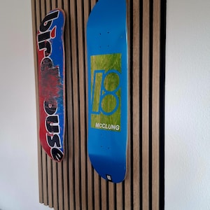 Pode incluir: Dois decks de skate montados num painel de parede com ripas de madeira verticais. O deck da esquerda é vermelho e azul com o texto birdhouse. O deck da direita é azul com um gráfico quadrado verde, o texto 18 e o nome MCCLUNG em branco.