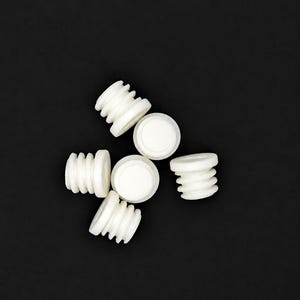 Op de afbeelding: Een verzameling van vijf kleine, witte, cilindrische plastic pluggen met een schroefdraadontwerp aan de buitenkant. Elke plug heeft een plat, rond bovenoppervlak en een geribbeld zijprofiel, gerangschikt in een groep.