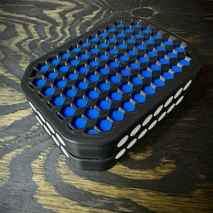 Op de afbeelding: Een zwarte rechthoekige container met een honingraatpatroon aan de bovenkant, met een blauw interieur. De zijkanten zijn geaccentueerd met witte ovale vormen. Het item staat op een houten oppervlak.