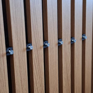 Könnte beinhalten: Eine Reihe vertikaler, hellbrauner Lamellen in Holzoptik, die parallel angeordnet sind. An der Seite jeder Lamelle befinden sich kleine schwarze Kunststoff-Montageclips, die jeweils mit einer silberfarbenen Metallschraube befestigt sind.