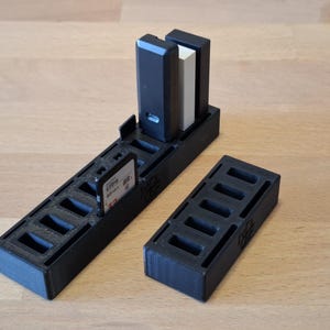 May include: Black 3D-printed storage organizer for SD cards and other small electronic devices. The organizer has multiple slots and compartments for storing various memory cards and accessories. Includes a separate, smaller storage unit.