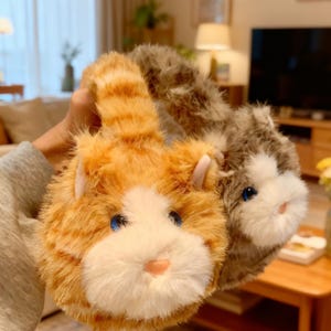 Könnte beinhalten: Zwei Ohrenschützer in Katzenform. Einer ist orange mit weißen und braunen Streifen, der andere grau und weiß. Beide haben blaue Augen und rosa Nasen. Die Ohrenschützer werden vor einem verschwommenen Hintergrund gehalten.