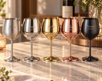 Puede incluir: Cinco copas de vino de acero inoxidable en acabados plata mate, plata pulida, oro, oro rosa y negro mate. Cada copa presenta un nombre grabado personalizado y un pequeño icono de corazón, con la copa negra mostrando además una fecha.
