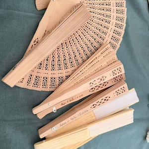 May include: Several open and closed wooden fans with intricate cut-out designs. Some fans have text that reads "Aiden & Katie 7.10.2026" and "Aiden & Katie 7.10.2026". The fans are light brown and beige in color.