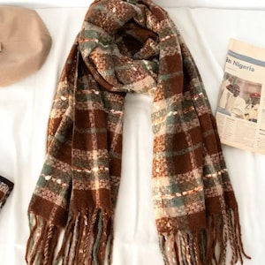 May include: A brown and green plaid scarf with a fringed edge. The scarf is rectangular and made of a soft, woven material. The plaid pattern features shades of brown, green, and white. A beige beret and a newspaper are in the background.