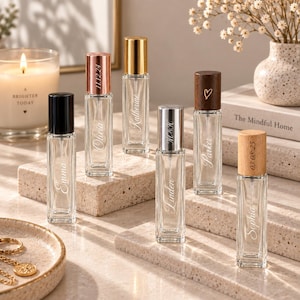 Puede incluir: Seis frascos de perfume de vidrio transparente con varios tapones de metal y madera. Cada frasco presenta un nombre o fecha grabados en la parte frontal. Los tapones incluyen acabados en negro, oro rosa, dorado, plateado, madera oscura y clara.