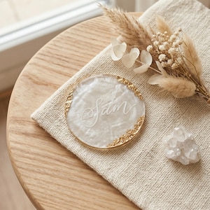 May include: A round, pearlescent coaster with gold leaf edging and the name "Sam" in elegant script. The coaster rests on a beige linen cloth, with dried flowers and a crystal cluster nearby. The coaster is a decorative item.