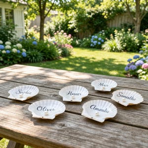 May include: Six white seashell place cards with names written in black script are arranged on a wooden table. Each shell has a small pearl. The table is outdoors with a garden in the background.