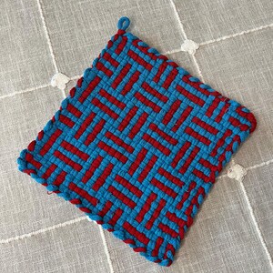 May include: A square, woven potholder in a vibrant pattern of red and turquoise blue. The potholder features a looped corner for hanging and a textured, geometric design. The edges are finished with a matching blue border.