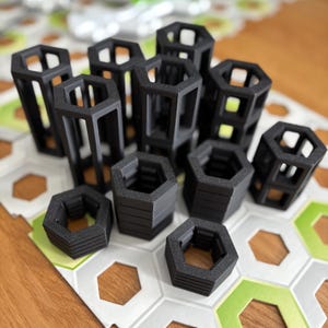 Puede incluir: Una colección de estructuras geométricas negras de marco abierto en varios tamaños. Algunas son altas y rectangulares, mientras que otras son más cortas y hexagonales. Están dispuestas sobre una superficie estampada con detalles en verde y blanco, sugiriendo un juego de construcción o rompecabezas.