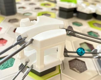 Puede incluir: Primer plano de un juego de circuito de canicas en progreso. La imagen presenta elementos arquitectónicos de plástico blanco, pistas metálicas y una canica de color turquesa. El tablero del juego tiene un diseño hexagonal.