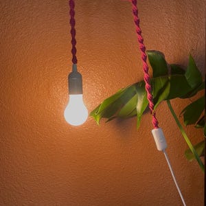 Puede incluir: Una lámpara colgante decorativa con un cordón de macramé rosa. La bombilla está encendida y se ve un interruptor blanco en el cable. La lámpara está suspendida contra una pared naranja texturizada, con una planta verde en el fondo.