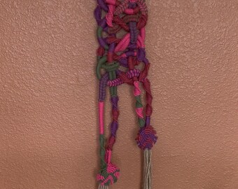 May include: A handmade macrame wall hanging featuring a metal ring at the top. The macrame design incorporates pink, purple, and green knotted cords, with two tassels and decorative flower-like accents at the bottom. The wall hanging is displayed against a peach-colored wall.