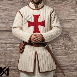 Può includere: Gambeson trapuntato color crema con un grande emblema a croce rossa sul petto. L'indumento medievale ha finiture marroni, un colletto alto e uno spacco frontale. Una cintura in pelle marrone, una tasca e una spada completano l'abito storico.