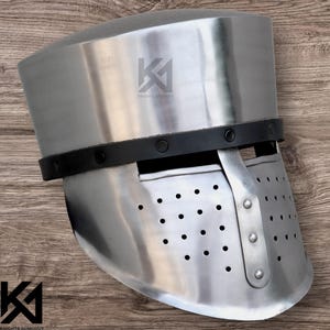 Könnte beinhalten: Ein polierter Stahlhelm mit einem genieteten schwarzen Lederband. Der Helm hat eine rechteckige Gesichtsplatte mit mehreren Belüftungslöchern. Der Text "KNIGHTS ARMOURY" ist links unten und rechts oben auf dem Helm zu sehen.