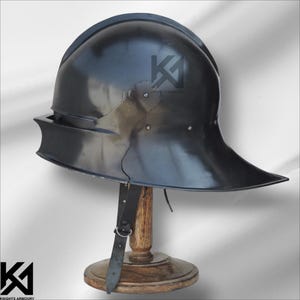 Op de afbeelding: Een zwarte metalen helm met een brede rand en een afgeronde bovenkant, tentoongesteld op een houten standaard. De helm heeft een glanzende afwerking en een leren riem. Het logo "KNIGHTS ARMOURY" is zichtbaar.