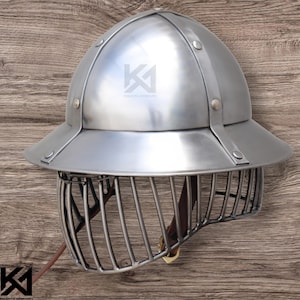 May include: A silver-colored helmet with a rounded top and a wide brim. The helmet features a cage-like face guard and is secured with rivets and straps. The text "KNIGHTS ARMOURY" is visible on the helmet.