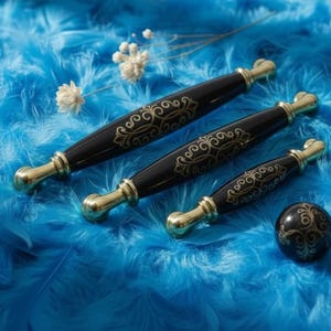 Può includere: Set di ferramenta decorativa per mobili nera e oro. Include tre maniglie di diverse dimensioni e un pomello rotondo, tutti con dettagli dorati ornati. Su uno sfondo di piume blu.