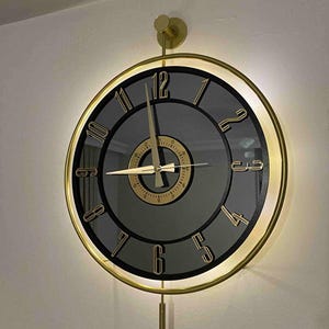 Może przedstawiać: Okrągły zegar ścienny ze złotą ramą, czarną tarczą i złotymi cyframi. Zegar ma złoty wahadło i jest zamontowany na białej ścianie. Wskazówki zegara są złote i jest podświetlany.