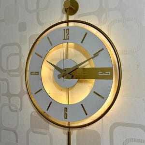 Peut inclure: Une horloge murale ronde avec un cadre doré et un cadran blanc. L'horloge présente des aiguilles dorées et des repères à 12, 3, 6 et 9. Elle possède un cercle intérieur lumineux et est fixée sur un mur à motifs. Le design de l'horloge est minimaliste.