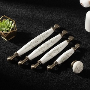 Può includere: Un set di cinque maniglie per mobili bianche e dorate e un pomello coordinato. Le maniglie hanno un centro in ceramica bianca con un motivo floreale e estremità in metallo dorato. È presente anche una piccola pianta grassa in una fioriera geometrica dorata.