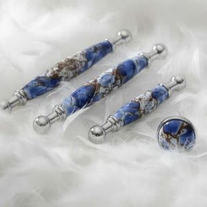 Pode incluir: Um conjunto de quatro peças de ferragens decorativas para armários. O conjunto inclui três puxadores e um botão. As ferragens apresentam um design marmorizado azul e castanho com detalhes metálicos prateados. As peças são exibidas numa superfície branca e plumosa.