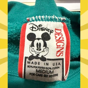 May include: Close-up of a teal sweatshirt collar with a Disney Designs tag. The tag features a smiling Mickey Mouse graphic and text that reads "MADE IN U.S.A., 50% POLYESTER/50% COTTON, MEDIUM, FOR CARE SEE REVERSE."