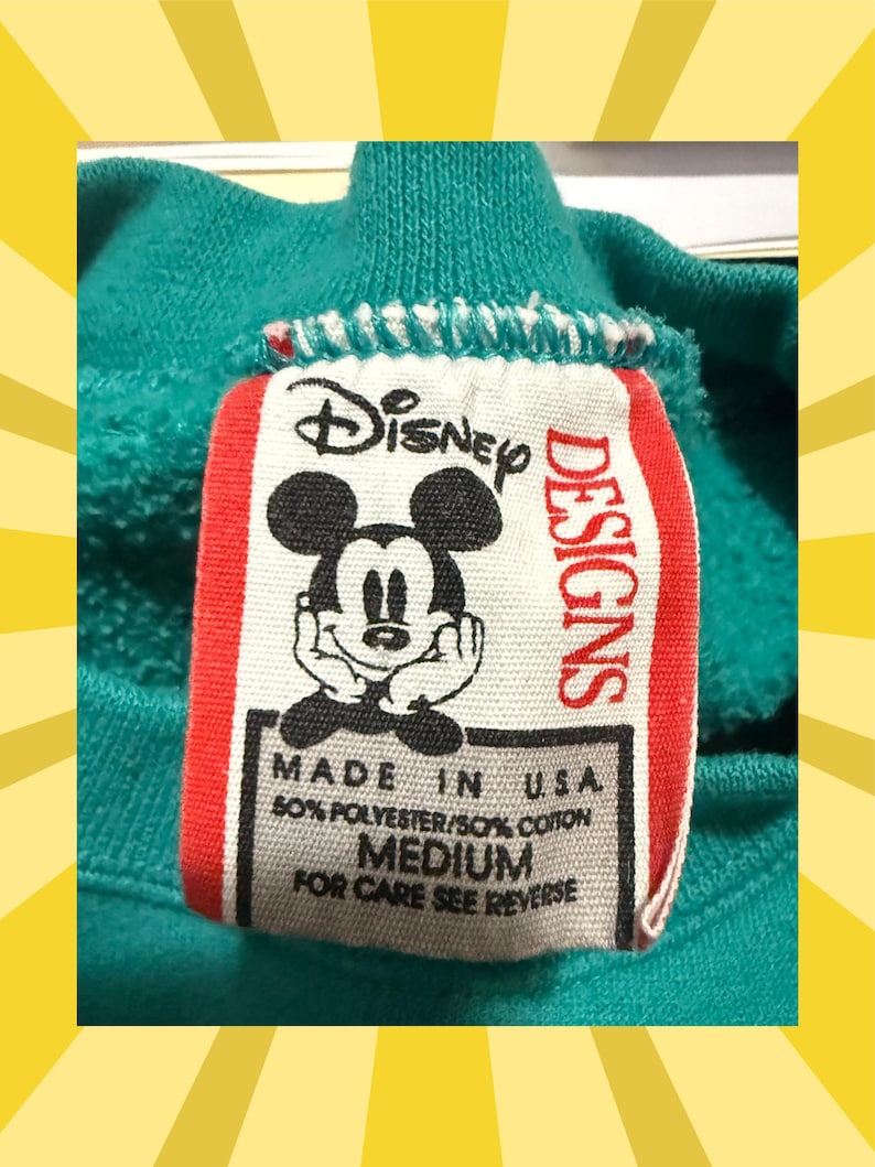 May include: Close-up of a teal sweatshirt collar with a Disney Designs tag. The tag features a smiling Mickey Mouse graphic and text that reads "MADE IN U.S.A., 50% POLYESTER/50% COTTON, MEDIUM, FOR CARE SEE REVERSE."