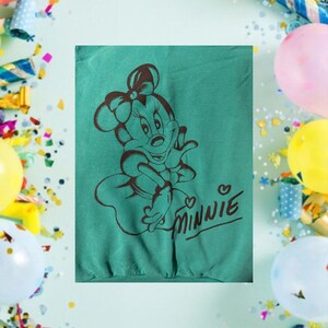 May include: Teal sweatshirt featuring a brown graphic of Minnie Mouse with a bow and the word "Minnie" in a cursive font. The design is centered on the shirt. The background includes balloons and confetti.