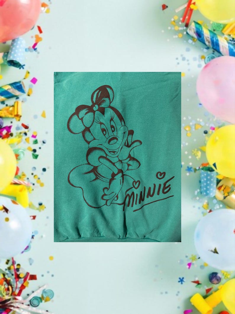 May include: Teal sweatshirt featuring a brown graphic of Minnie Mouse with a bow and the word "Minnie" in a cursive font. The design is centered on the shirt. The background includes balloons and confetti.