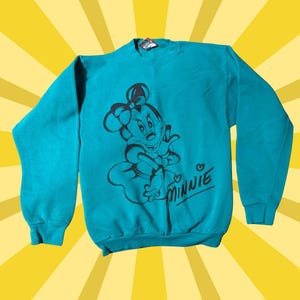 May include: Teal sweatshirt featuring a black graphic of Minnie Mouse with a bow, dress, and the word "Minnie" in cursive. The sweatshirt has long sleeves and a crew neck. The background is yellow with radial lines.