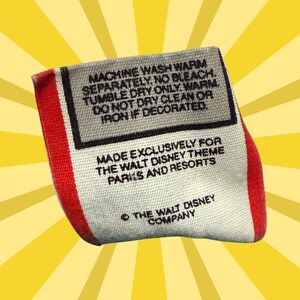 May include: A rectangular tag with red and white edges, featuring black text on a gray background. The tag includes washing instructions and states it was made exclusively for Walt Disney Theme Parks and Resorts. The Walt Disney Company copyright is also visible.