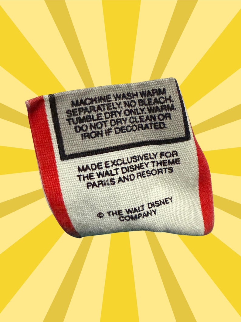 May include: A rectangular tag with red and white edges, featuring black text on a gray background. The tag includes washing instructions and states it was made exclusively for Walt Disney Theme Parks and Resorts. The Walt Disney Company copyright is also visible.